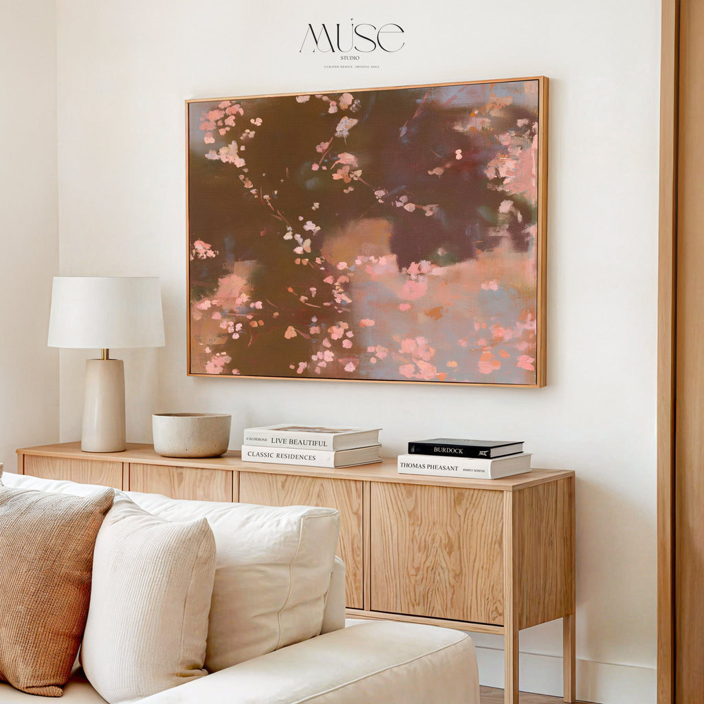 A framed abstract floral painting with pink blossoms and dark brown background hangs on a cream wall above a light wood console table in a styled living room. The brand name 'MUSE DESIGN' is printed above the artwork.
