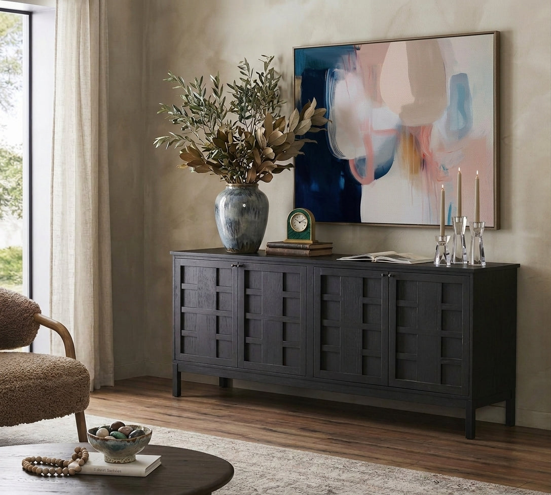 A dark charcoal gray wooden sideboard cabinet styled in a warm, modern living room setting with a large abstract painting, a blue-gray ceramic vase with dried leaves, and decorative accents.
