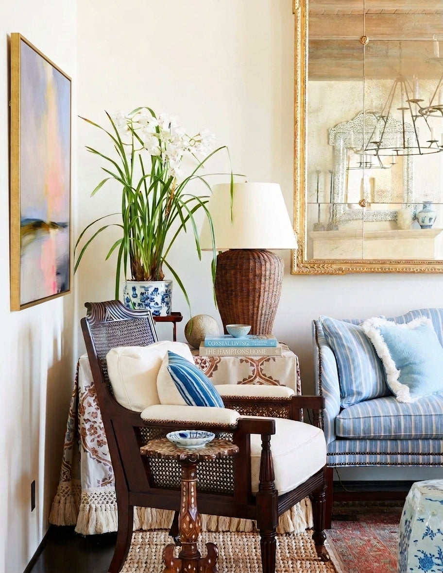 A framed vertical abstract impressionistic painting with soft strokes of blue, pink, and yellow hangs on a light wall in a sophisticated coastal-style room. Below the artwork is a dark, carved wood armchair with an white cushion and blue and white striped pillow, next to a table with a woven lamp and large white orchid.