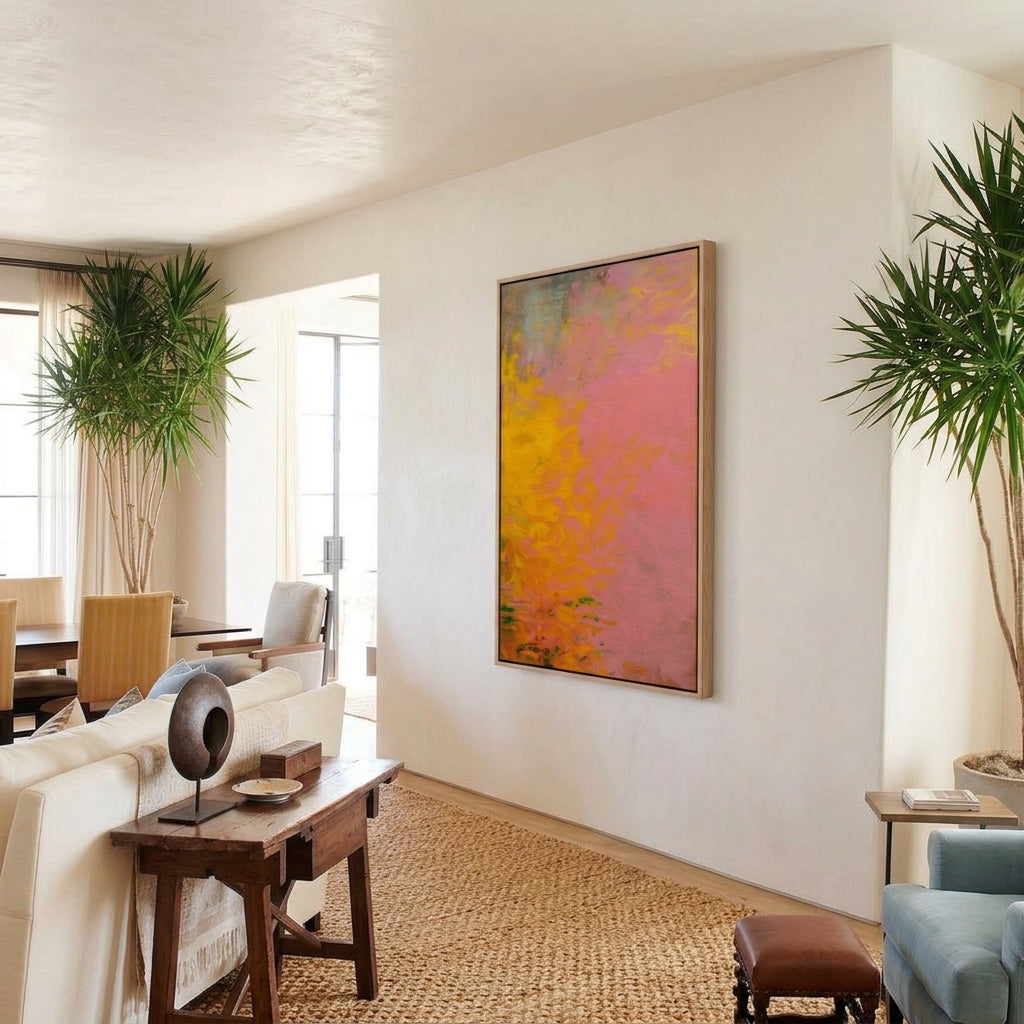 Framed pink and yellow abstract painting canvas hung on a plaster wall in a warm living room staged with indoor plants, jute rug, and modern furniture.