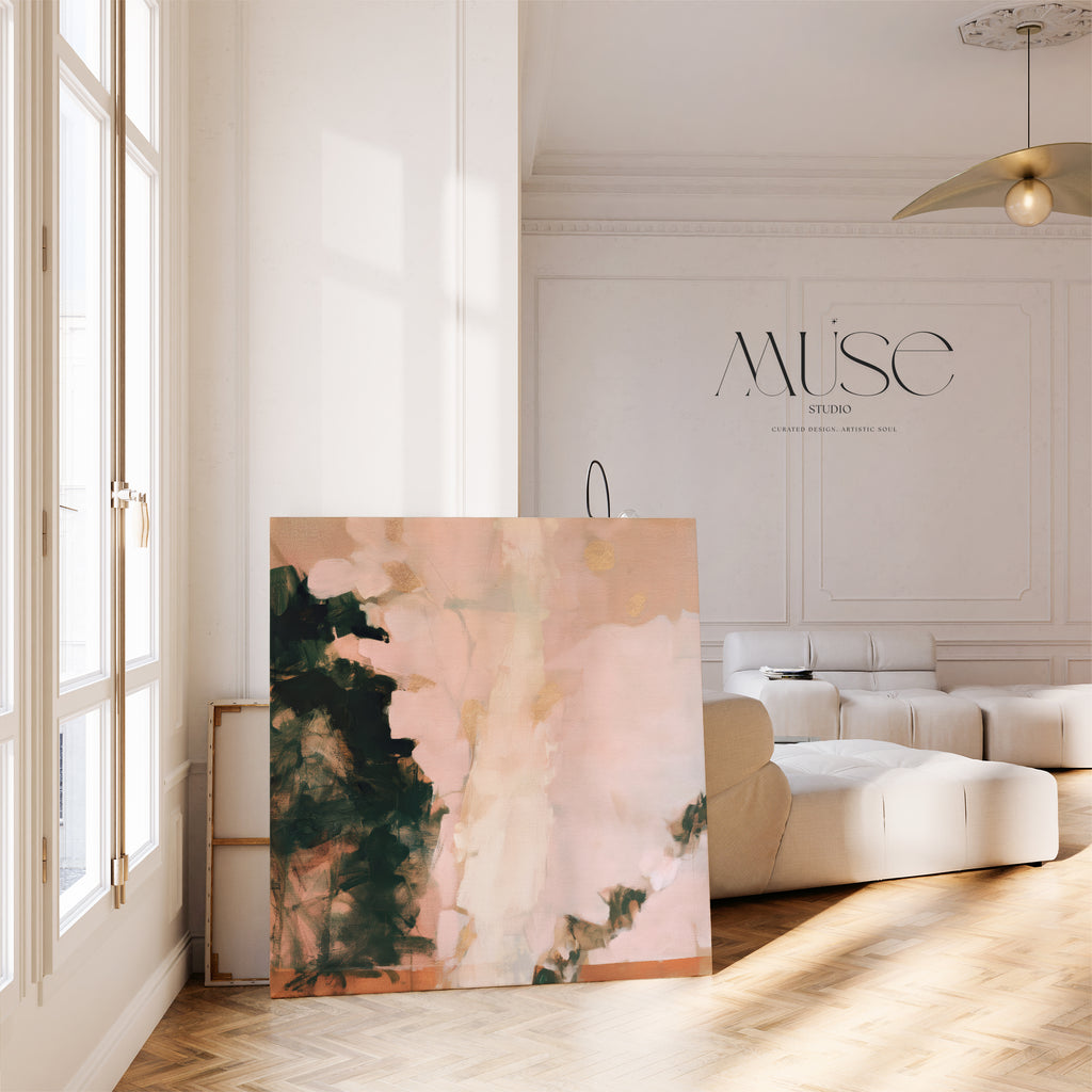 A large square abstract canvas painting titled "Morning Blush No. 1181" resting on a light chevron wood floor inside a sunlit, modern living room with tall French windows, a white modular sofa, and high molded ceilings. The artwork showcases soft pink blush hues contrasted with dark forest green and subtle metallic gold brushstrokes.