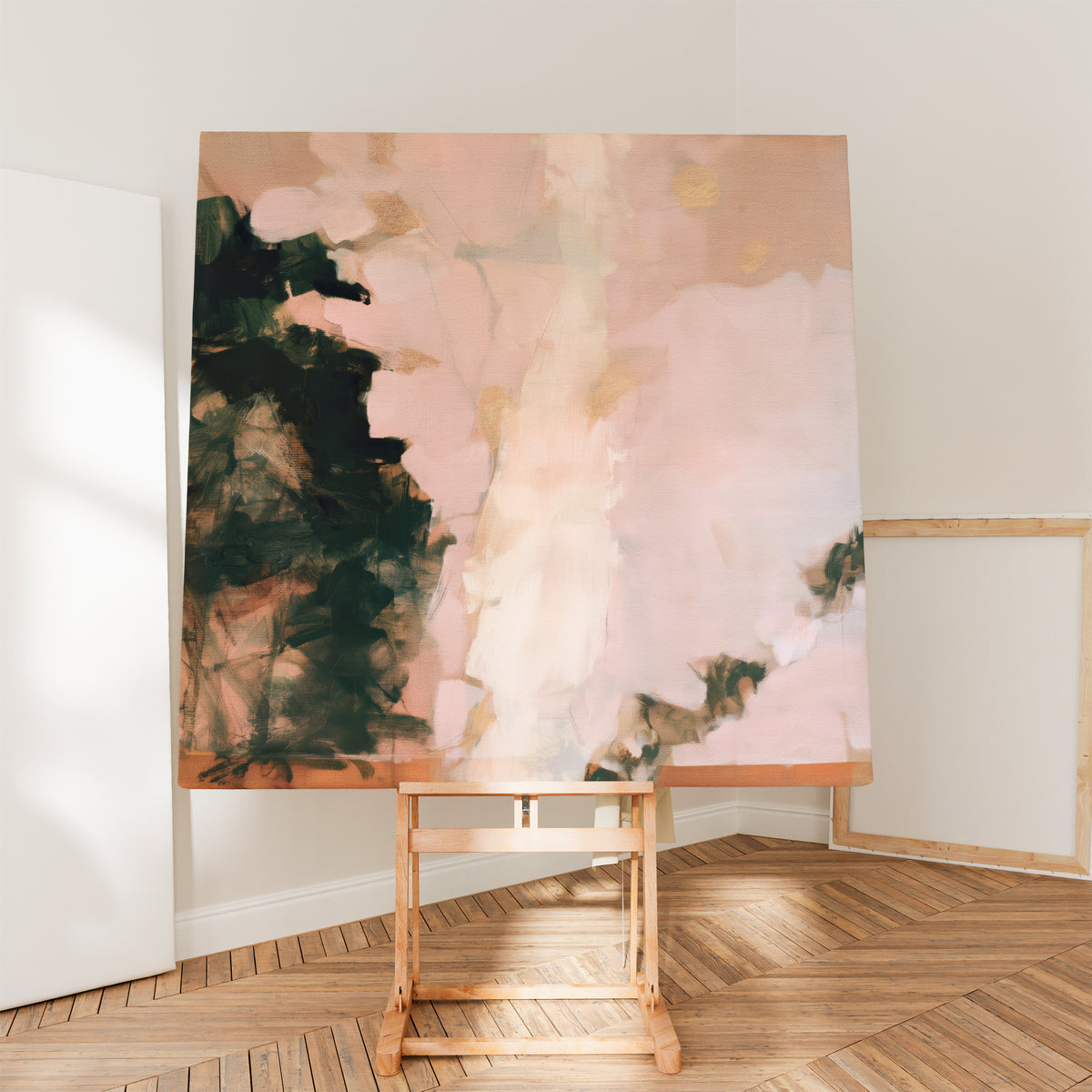A large square canvas featuring pink, green, and gold abstract brushwork presented on a light-wood studio easel set over herringbone parquet flooring in a bright art studio corner.