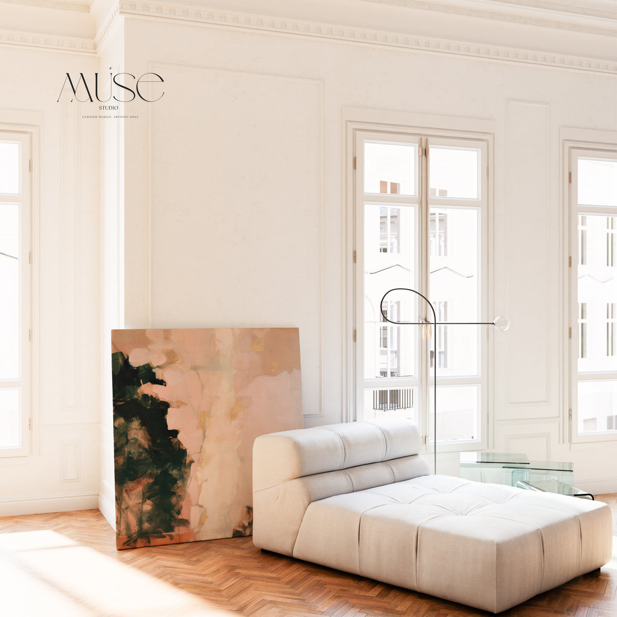 A oversized square canvas art print leaning against a white wall next to a low-profile tufted white lounge chair in a bright room with classic wall paneling and tall double windows.