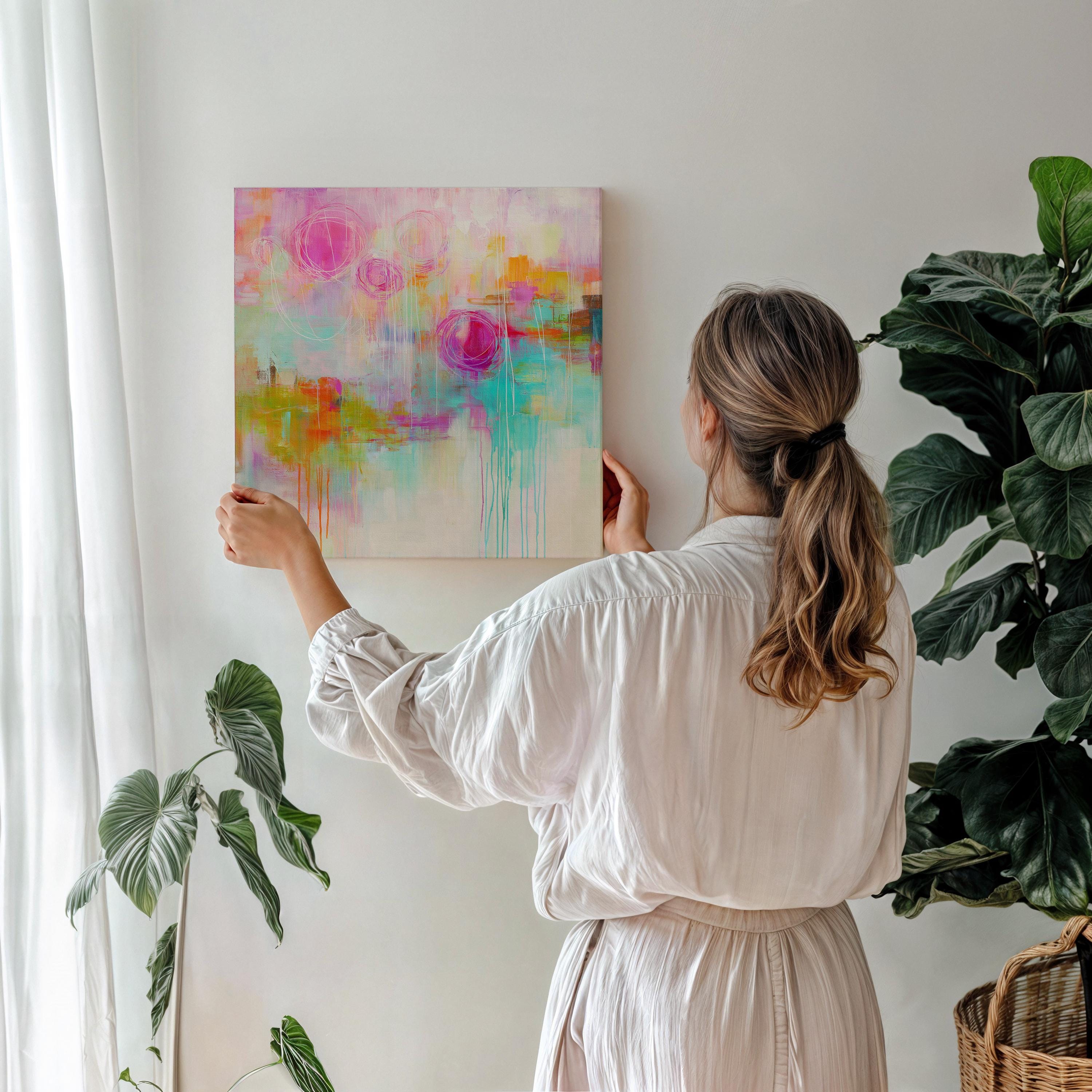 Large abstract canvas art in bright pink, aqua, orange, and yellow tones with loose circular lines and painterly drips. Staged in a sunlit boho-modern interior, this artwork evokes creative energy and bold eclectic style.