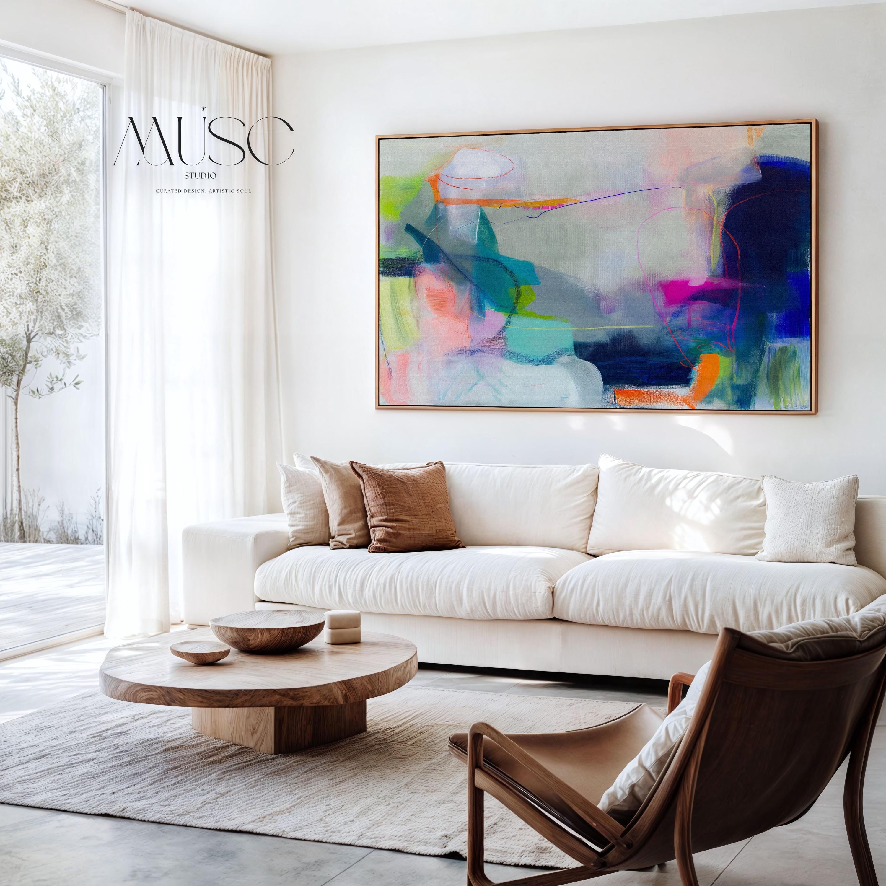 Large horizontal abstract wall art in bright blue, neon pink, teal, and multicolor tones displayed above a white sofa in a modern neutral living room with natural light and minimal decor. Vibrant color pop artwork available as framed canvas or fine art print by Muse Studio.