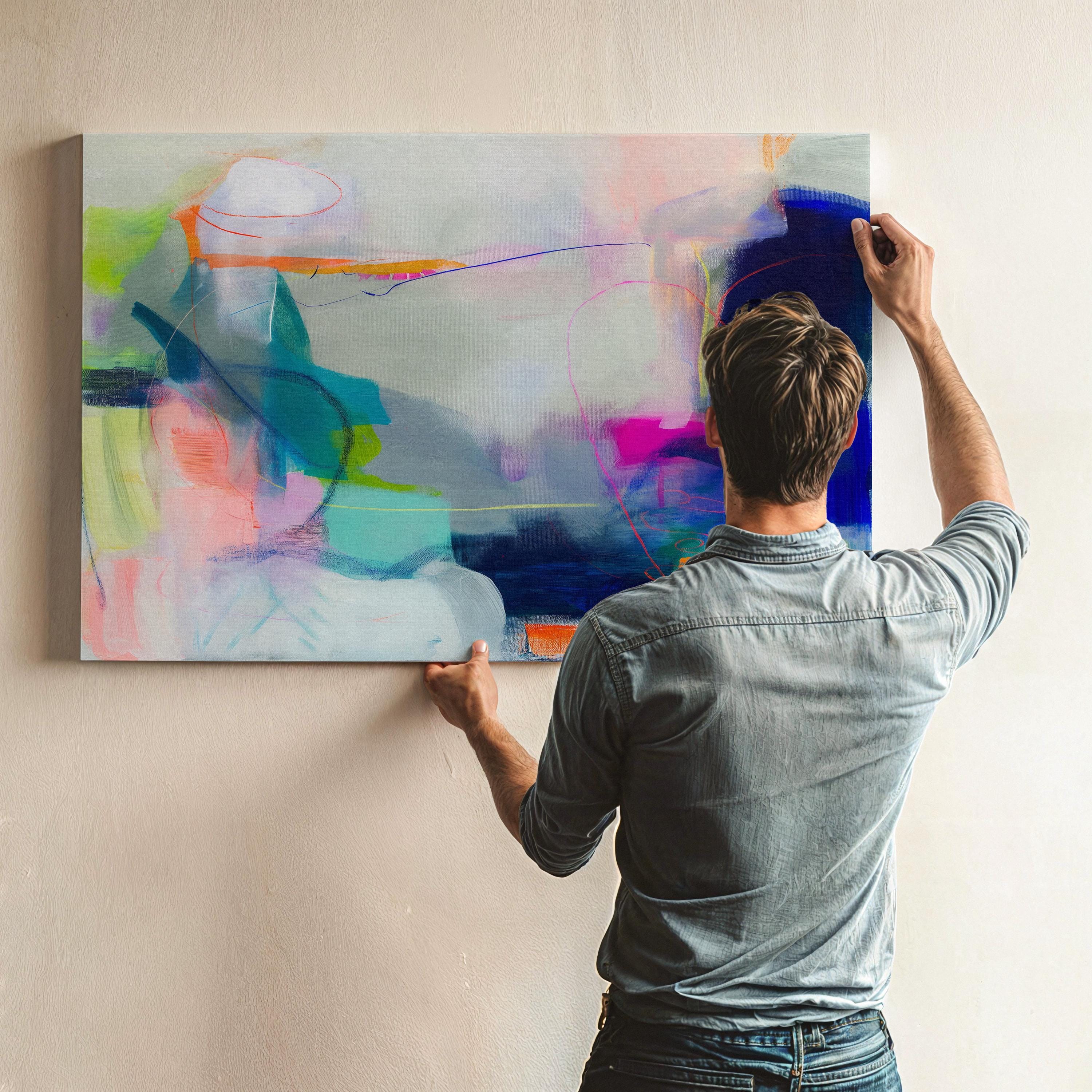 Large horizontal abstract wall art in bright blue, neon pink, teal, and multicolor tones displayed above a white sofa in a modern neutral living room with natural light and minimal decor. Vibrant color pop artwork available as framed canvas or fine art print by Muse Studio.
