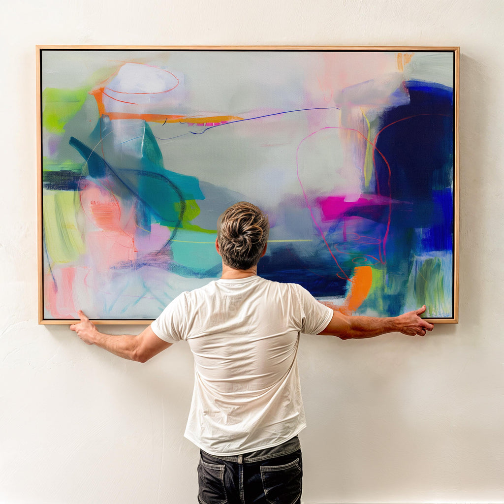 Large horizontal abstract wall art in bright blue, neon pink, teal, and multicolor tones displayed above a white sofa in a modern neutral living room with natural light and minimal decor. Vibrant color pop artwork available as framed canvas or fine art print by Muse Studio.