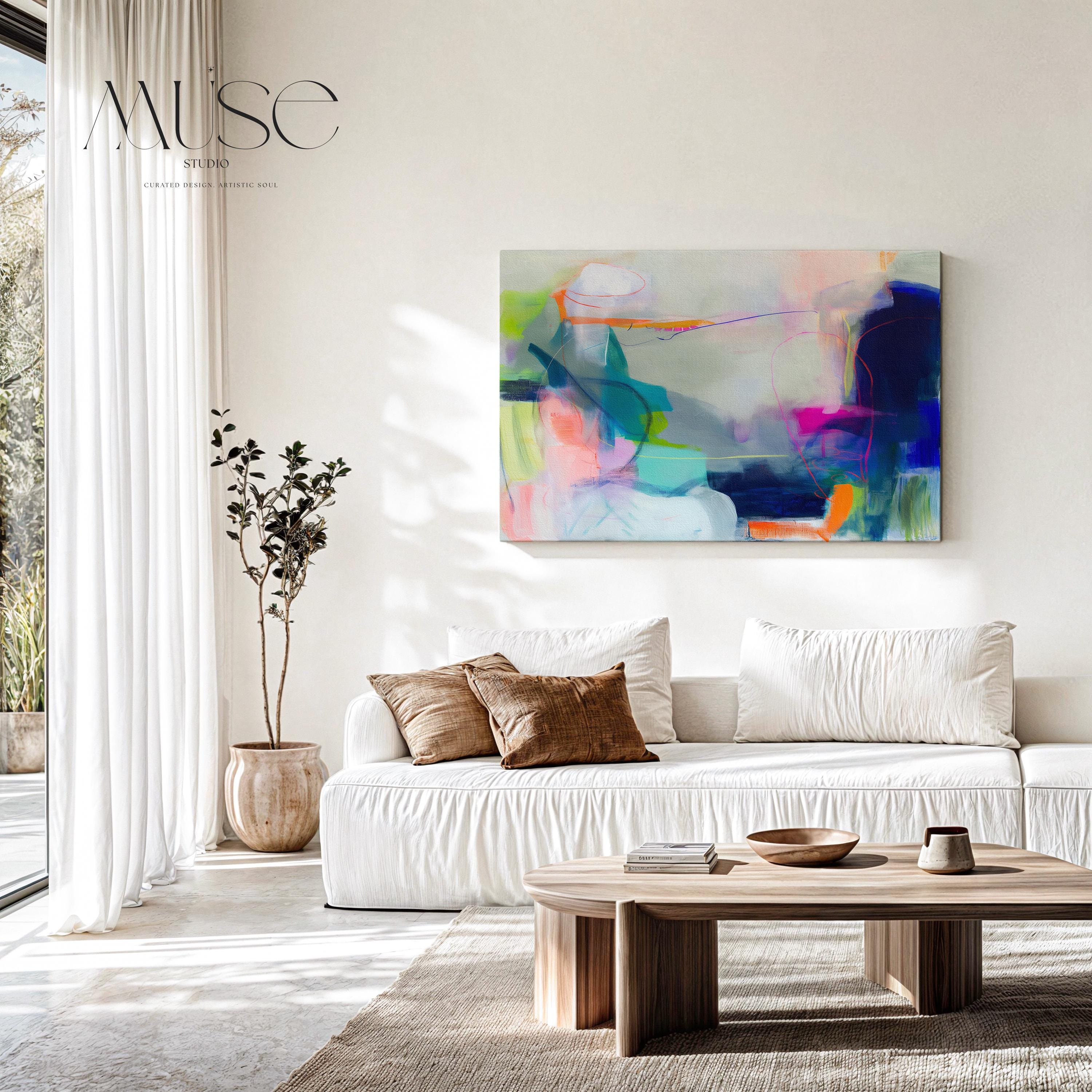 Large horizontal abstract wall art in bright blue, neon pink, teal, and multicolor tones displayed above a white sofa in a modern neutral living room with natural light and minimal decor. Vibrant color pop artwork available as framed canvas or fine art print by Muse Studio.