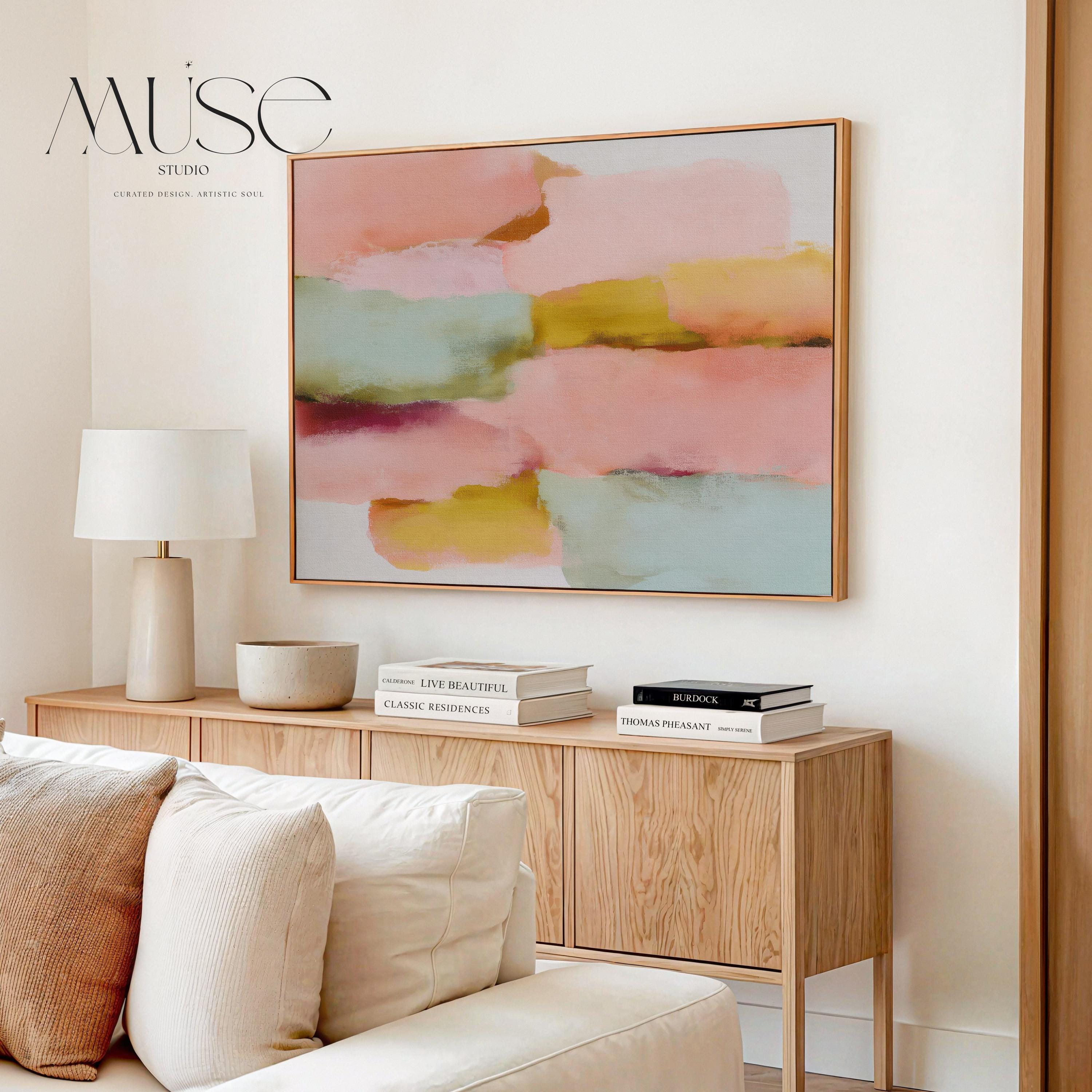 Large horizontal abstract framed canvas wall art featuring blush pink, coral, muted aqua blue, golden ochre, and plum layered brushstrokes, styled above a neutral sofa in a modern living room, offering warm contemporary color and balanced movement for oversized wall placement.