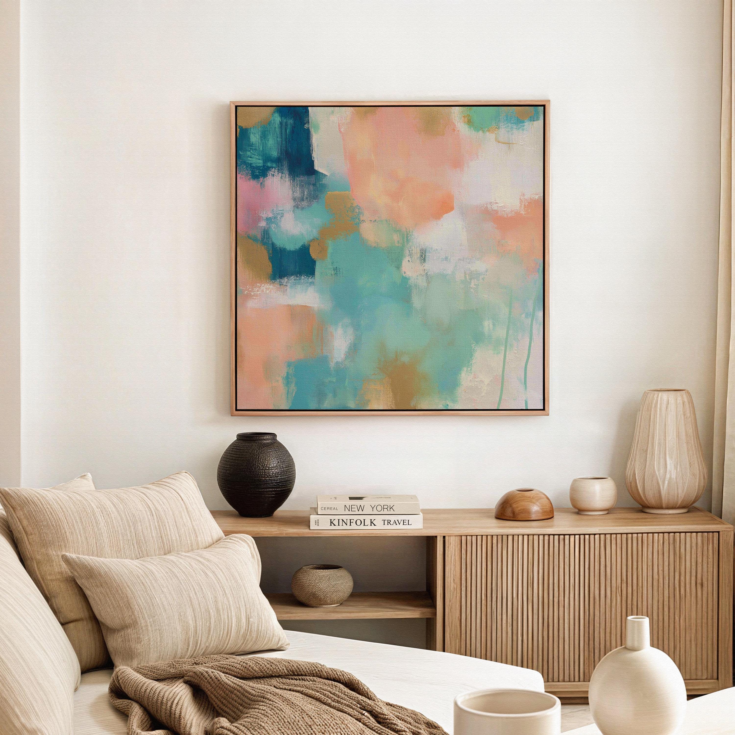 Large square abstract framed canvas wall art featuring coral, peach, teal, aqua blue, and warm sand tones layered over a soft cream background, styled above a neutral sofa, adding soft color contrast and airy painterly texture to a curated living space.