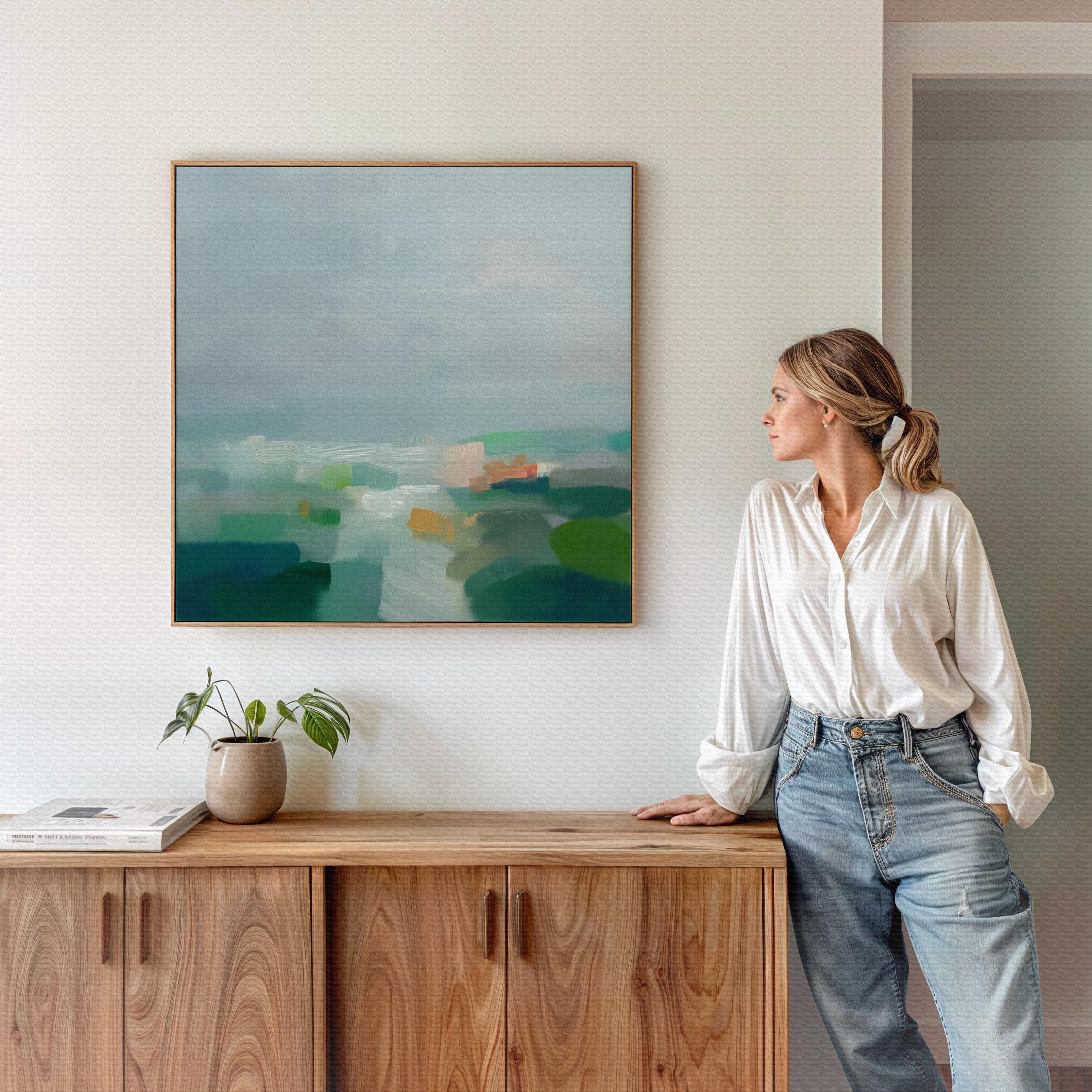 Large square abstract decor wall art featuring mist blue, sea glass green, and soft teal tones, framed in natural wood and styled in a calm contemporary living room, creating a serene coastal-inspired focal point for spacious interiors.