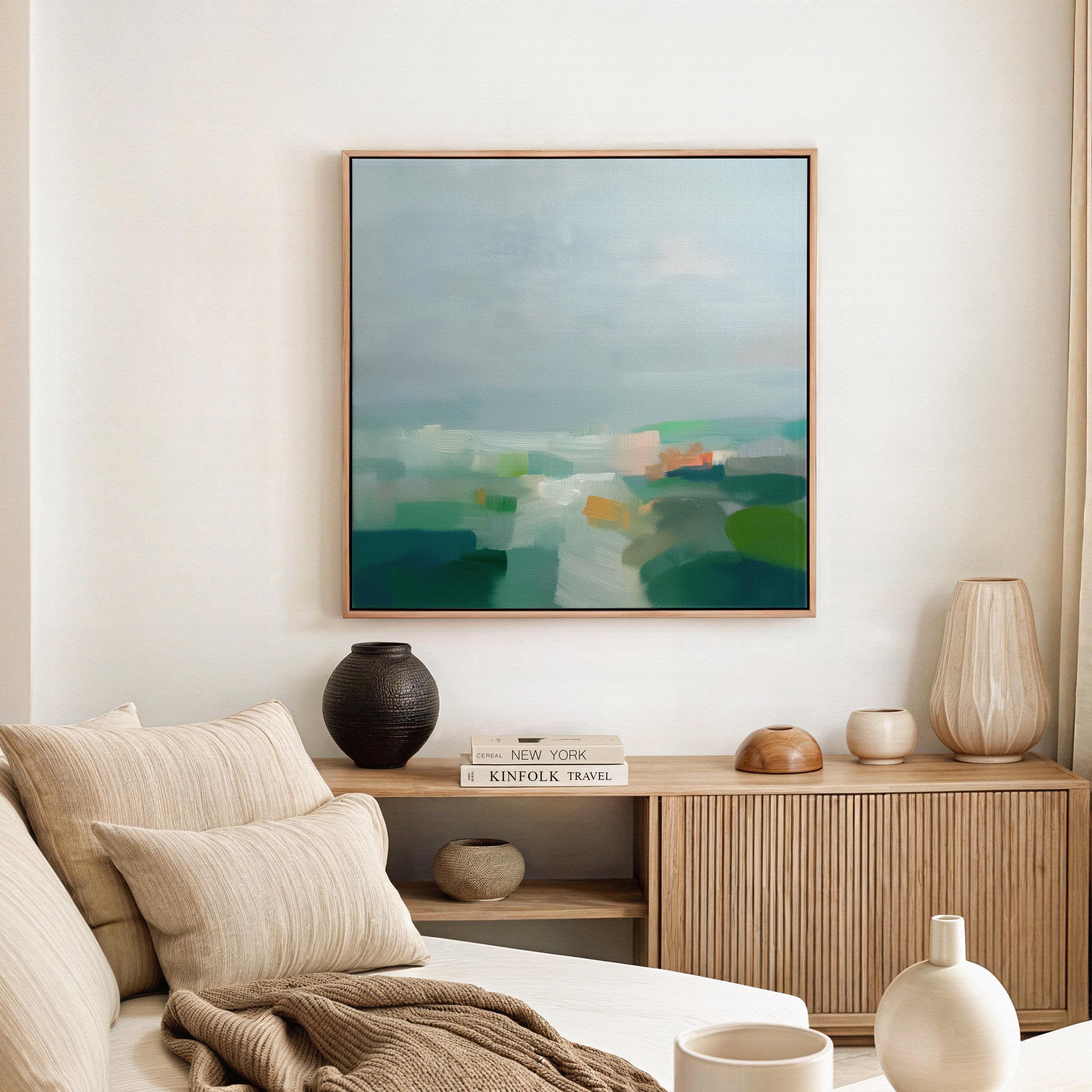 Large square framed canvas wall art in sage green, soft blue, and muted cream tones styled in a warm neutral farmhouse interior, creating a calm countryside-inspired focal point with soft horizon depth.