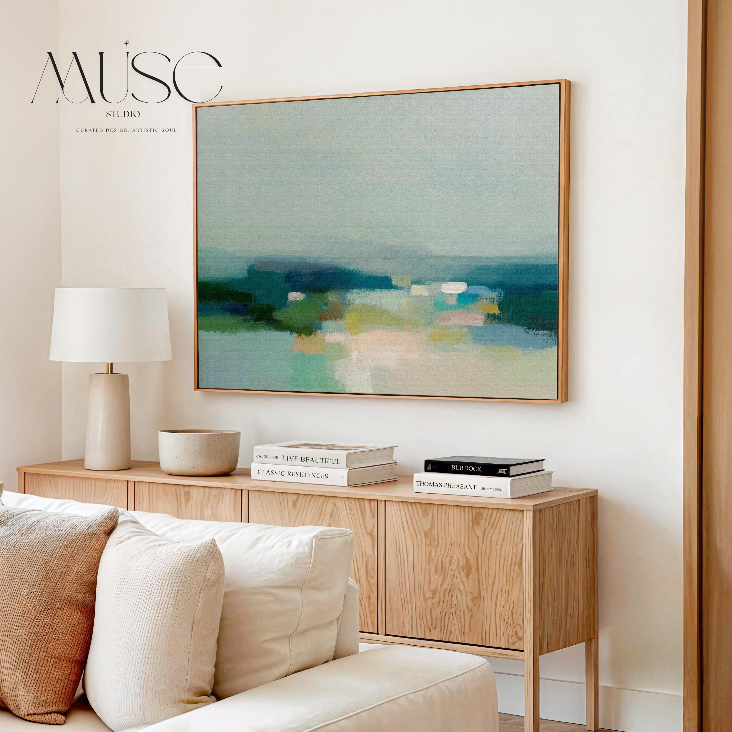 Large horizontal framed canvas wall art in sage green, deep teal, cream, and soft blue tones styled above a light wood console in a warm neutral interior, creating a calm landscape-inspired focal point.