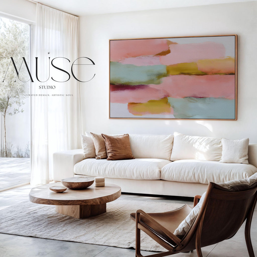 Large horizontal abstract framed canvas wall art featuring blush pink, coral, muted aqua blue, golden ochre, and plum layered brushstrokes, styled above a neutral sofa in a modern living room, offering warm contemporary color and balanced movement for oversized wall placement.
