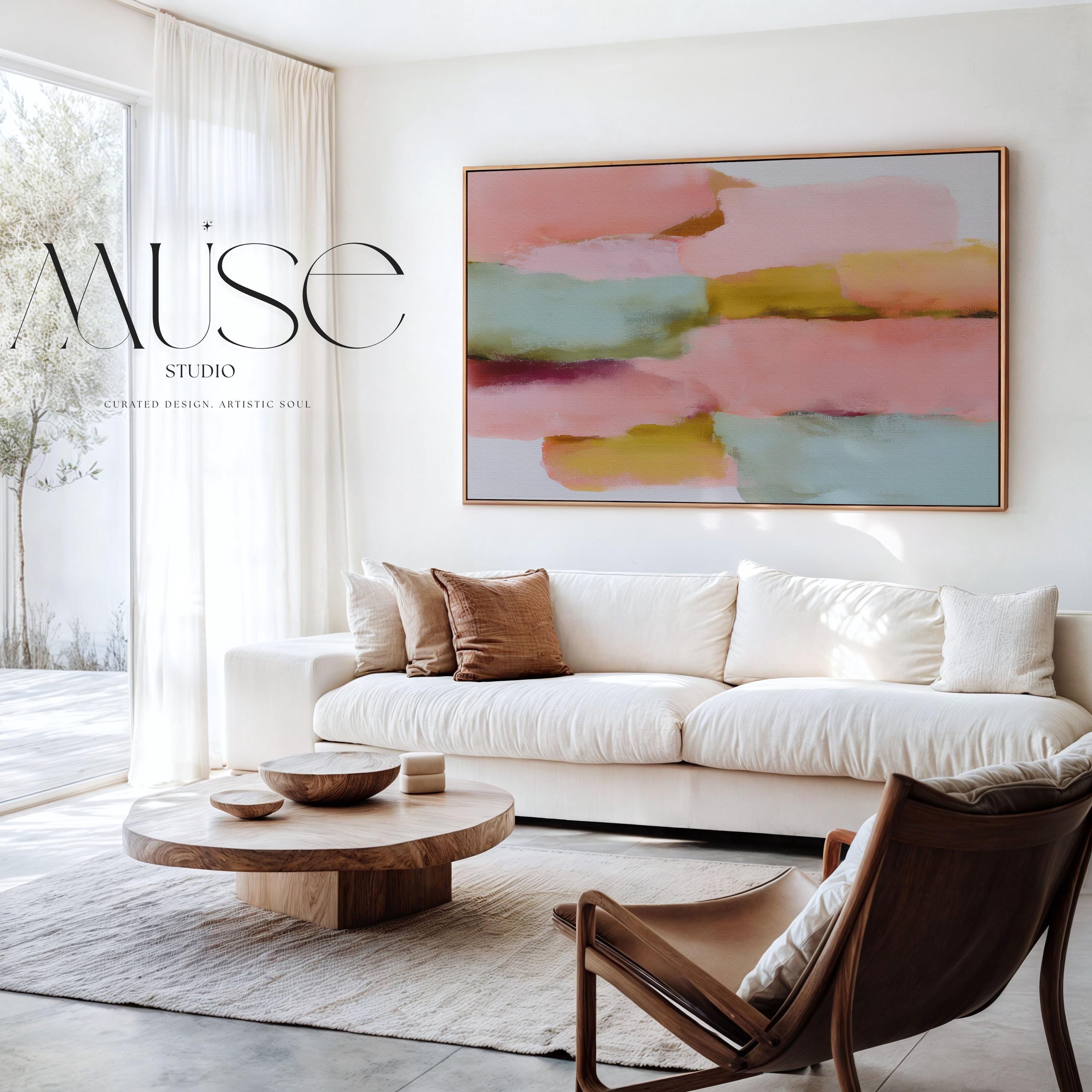 Large horizontal abstract framed canvas wall art featuring blush pink, coral, muted aqua blue, golden ochre, and plum layered brushstrokes, styled above a neutral sofa in a modern living room, offering warm contemporary color and balanced movement for oversized wall placement.