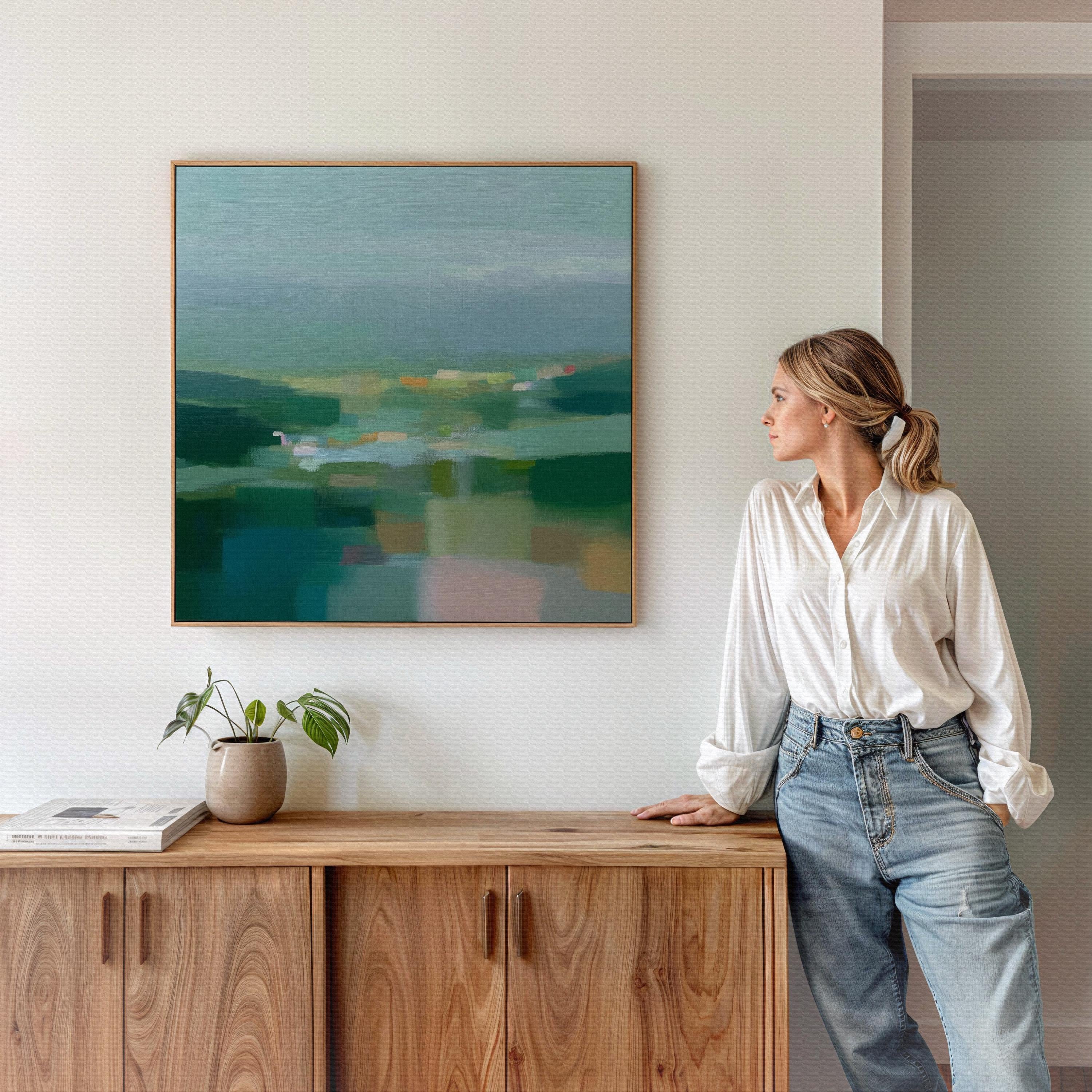 Large square framed canvas wall art in layered sage, deep green, and misty blue tones styled against a light neutral wall, creating a calm nature-inspired focal point in a refined interior.