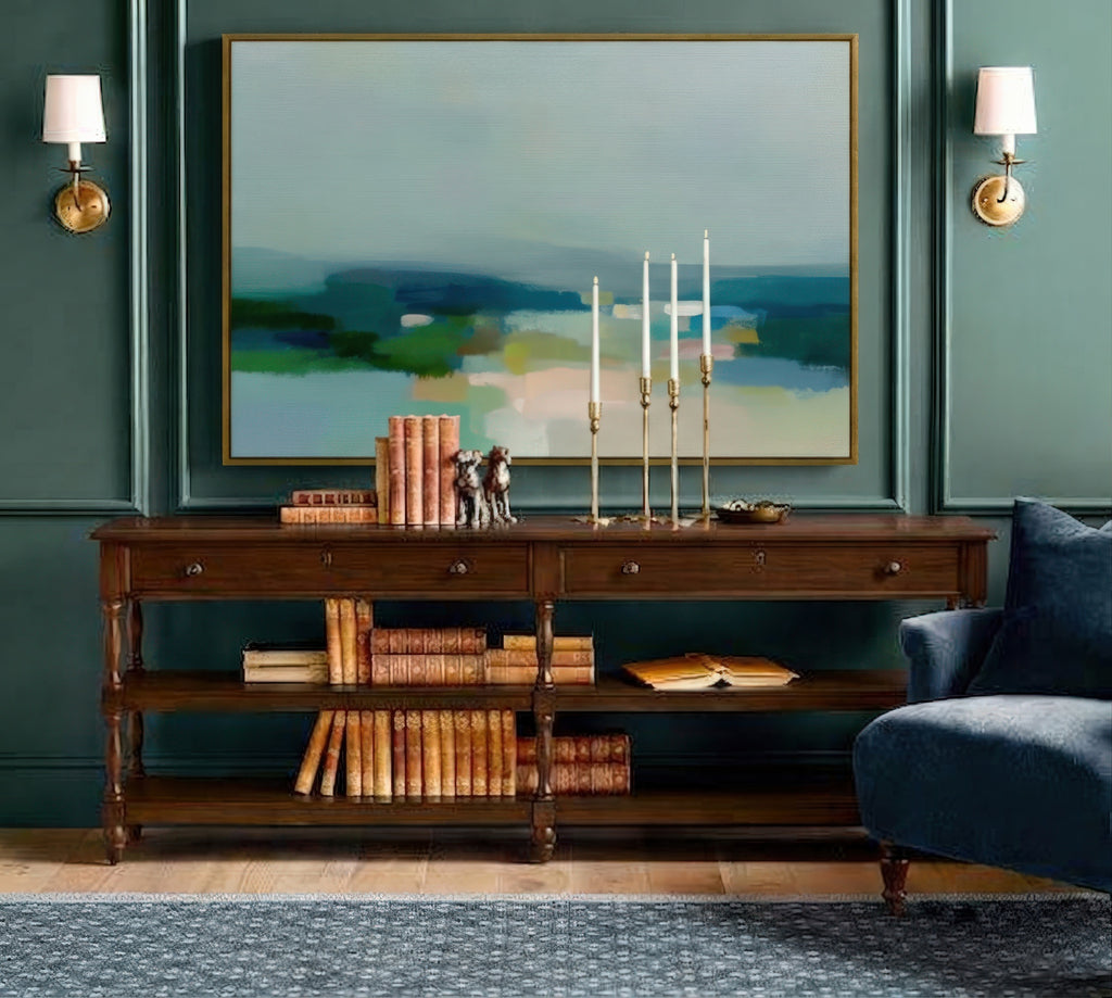 Nestled against a dramatic teal paneled wall, a rich dark wood console table commands attention. Above it hangs a large abstract landscape, its muted greens and blues evoking the tranquil colors of nature.  Brass sconces frame the art, casting a warm glow, while below, a collection of vintage books and elegant brass candlesticks complete this scene of sophisticated, classic style.